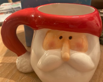 Puede incluir: Una taza de cerámica de Papá Noel con gorro y asa rojos, barba blanca y mejillas rosadas. La taza está diseñada para parecerse a la cara de Papá Noel, perfecta para bebidas navideñas. La taza está sobre una superficie de madera.