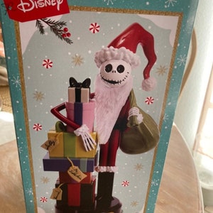 May include: A Christmas nutcracker featuring Jack Skellington from The Nightmare Before Christmas. He is dressed as Santa Claus and holding a bag of gifts. The nutcracker is on a base with the text "Jack Skellington" and "Nutcracker Cusse Noisette".