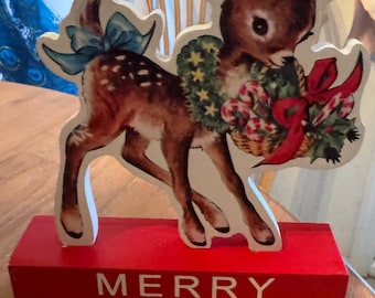 Puede incluir: Decoración navideña de madera con un ciervo con una corona y una cesta de bastones de caramelo. El ciervo es marrón con detalles blancos y un lazo azul. La base roja tiene la inscripción "MERRY CHRISTMAS" en blanco.