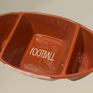May include: An orange, football-shaped serving dish with three compartments. The word "FOOTBALL" is written in white on the central section. The dish's design is inspired by a football, perfect for serving snacks.
