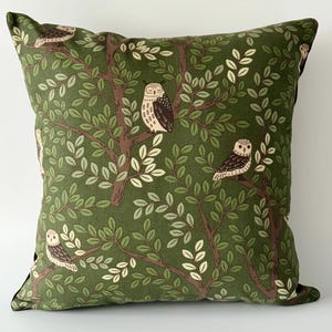 May include: Square cushion with a green background featuring a tree design with owls perched on branches. The cushion has a dark green border and a repeating pattern of owls and leaves in shades of brown, green, and cream.