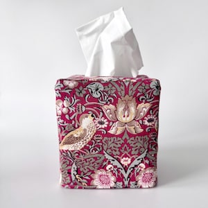 May include: A square tissue box cover featuring a vibrant magenta background with a detailed woven pattern of beige birds, grey foliage, and pink floral motifs. A single white tissue is pulled through the top opening of the fabric box cover.