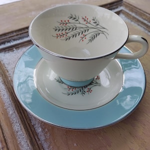 May include: A vintage teacup and saucer set with a delicate floral design. The cup is white with a silver rim and features a sprig of red berries and gray leaves. The saucer is light blue with a silver rim and a matching floral design.
