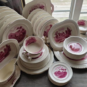 May include: A collection of antique white porcelain dinnerware with a red landscape design. The set includes plates, bowls, cups, saucers, and serving dishes. The edges of the plates and dishes have a beaded trim.