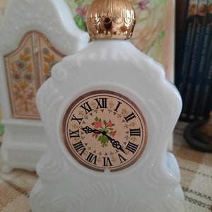May include: White porcelain clock shaped bottle with a gold top and a clock face with Roman numerals and a floral design.
