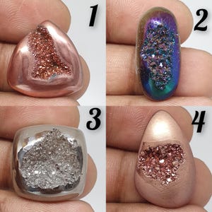 May include: Four polished gemstone cabochons. The first is a triangular shape with a copper-coloured exterior and a sparkling, reddish-brown interior. The second is an oval shape with a shimmering, iridescent surface. The third is a square shape with a silver interior. The fourth is a teardrop shape with a copper exterior and a reddish-brown interior.