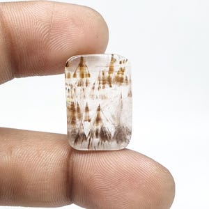 May include: A rectangular gemstone with a clear base and intricate brown and gold inclusions. The inclusions create a pattern of vertical lines and shapes, resembling a landscape. The stone is held between fingers against a white background.