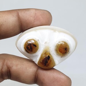 May include: A smooth, white, triangular-shaped stone cabochon featuring three distinct, embedded amber-colored circular and triangular inclusions with dark brown, branch-like patterns visible within the translucent, polished surface.