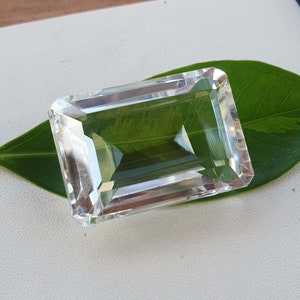 May include: A clear, rectangular, faceted gemstone with sharp edges and a polished surface. The stone is transparent, allowing light to pass through its geometric structure, and it is positioned directly on top of a single, smooth, green leaf.