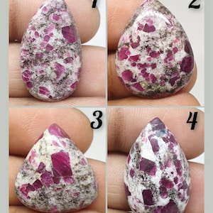 May include: Four close-up images of ruby gemstones. Each stone has a teardrop or irregular shape, with a mix of deep pink and white colors. The stones are held against a neutral background, showcasing their natural textures and variations.