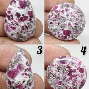 以下が含まれることがあります： さまざまな色の4つの宝石。2つは涙の形、2つは丸いです。宝石は白いベースに濃いピンクと濃い灰色のインクルージョンがあります。宝石は手で持たれています。