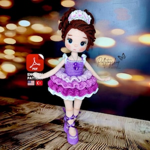 May include: Crochet doll wearing a purple and white dress with a crown and a necklace. The doll is standing on a wooden surface.