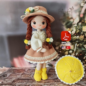 Puede incluir: Muñeca de crochet con cabello castaño que lleva una camisa blanca, una falda marrón y botas amarillas. La muñeca sostiene un paraguas amarillo de crochet. La muñeca lleva un sombrero de crochet marrón y blanco con flores amarillas. La imagen incluye el texto "PDF ENGLISH PATTERN".