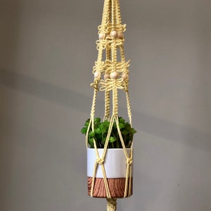 May include: A yellow macrame plant hanger with wooden beads and a white planter with a brown wood-grain pattern. The hanger is hanging from a hook on a light grey wall.