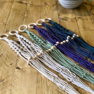 May include: Five macrame plant hangers in different colours, including white, green, purple, and blue. Each hanger has wooden beads and a wooden ring at the top.