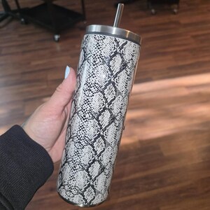 Puede incluir: Un vaso de acero inoxidable con un patrón de piel de serpiente blanco y negro. El vaso tiene una tapa y una pajita.