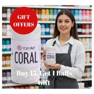 Könnte beinhalten: Eine Frau hält zwei Spulen YarnArt Coral-Garn. Die große Spule ist weiß mit einem Etikett mit der Aufschrift "CORAL" in großen schwarzen Buchstaben. Ein roter Kreis darüber zeigt "GESCHENKANGEBOTE". Der Hintergrund ist ein Regal voller farbenfroher Garne.