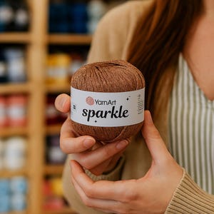 Op de afbeelding: Een bol bruin garen met een wit etiket met de tekst "YarnArt sparkle". Het garen wordt door iemand in de handen gehouden. De achtergrond toont schappen met garen.