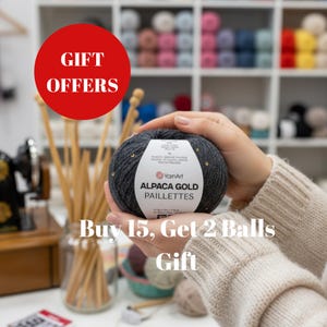 Op de afbeelding: Een donkergrijze bol Alpaca Gold Paillettes garen met gouden pailletten, vastgehouden in handen. Het label luidt "YarnArt". Een rode cirkel met witte tekst zegt "GIFT OFFERS". Tekst hieronder luidt "Buy 15, Get 2 Balls Gift".