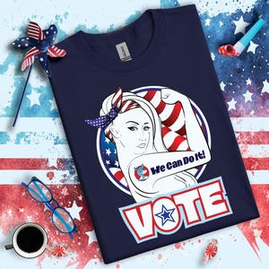 May include: Navy blue t-shirt with a graphic of a woman with a red, white, and blue bandana and a donkey tattoo on her arm. The graphic is in a circle with the text "We Can Do It!" and "VOTE" below the circle.