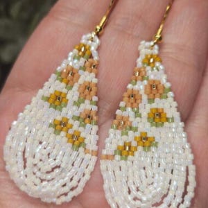 May include: Teardrop-shaped beaded earrings featuring a floral pattern in white, peach, and yellow glass beads. The design includes small green accents and gold-toned metal ear wires. The beads have an iridescent finish, creating a subtle shimmer.