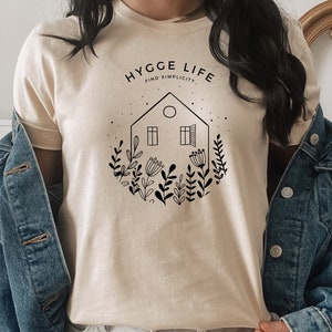May include: A beige t-shirt featuring a black graphic design of a house with windows and a circle in the roof, surrounded by flowers and plants. The text "HYGGE LIFE FIND SIMPLICITY" is above the house.