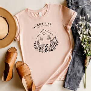 May include: A pink t-shirt featuring a black graphic design of a house with windows and flowers. The text "HYGGE LIFE FIND SIMPLICITY" is printed on the shirt.