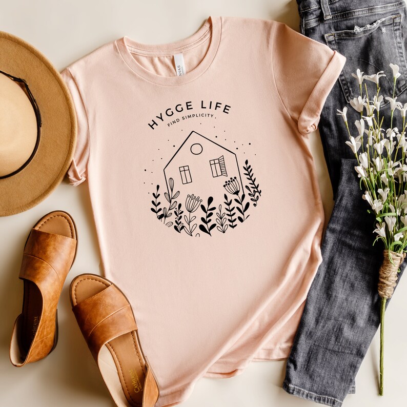 May include: A pink t-shirt featuring a black graphic design of a house with windows and flowers. The text "HYGGE LIFE FIND SIMPLICITY" is printed on the shirt.