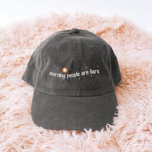 May include: A dark grey corduroy baseball cap with the embroidered text "morning people are liars" in white, with a sun graphic. The cap is displayed on a peach-coloured, fluffy surface.