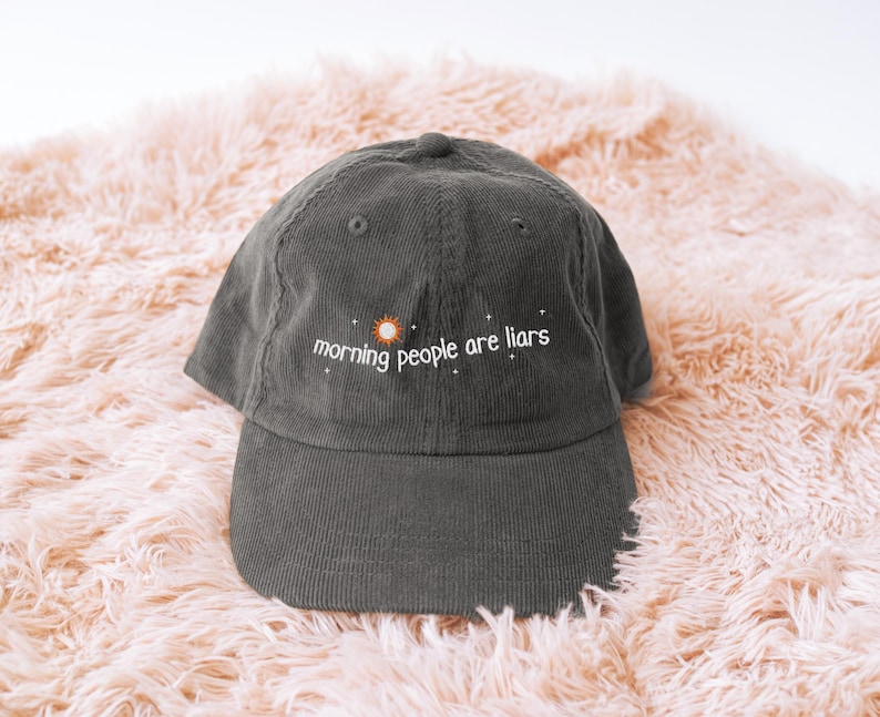May include: A dark grey corduroy baseball cap with the embroidered text "morning people are liars" in white, with a sun graphic. The cap is displayed on a peach-coloured, fluffy surface.
