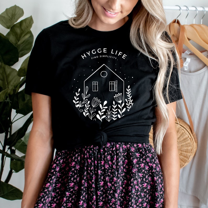 May include: A black t-shirt featuring a white graphic design of a house with a window and a door, surrounded by flowers and plants. The text "HYGGE LIFE FIND SIMPLICITY" is above the house.