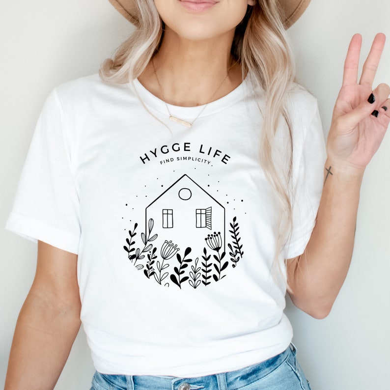 May include: White t-shirt featuring a black and white graphic design of a house with windows and a door. The graphic also includes flowers and leaves. The text "Hygge Life Find Simplicity" is above the house.