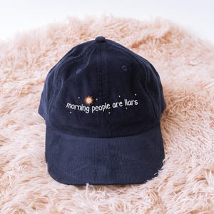May include: A navy blue corduroy baseball cap featuring the embroidered phrase "morning people are liars" with a sun and star design. The cap has a curved peak and a button on top. It is displayed on a peach-coloured, fluffy surface.