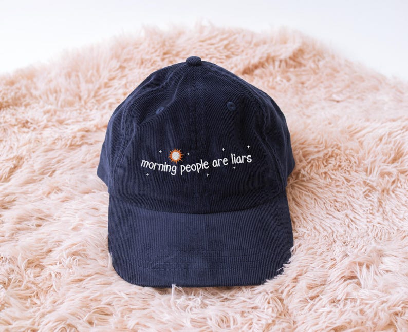 May include: A navy blue corduroy baseball cap featuring the embroidered phrase "morning people are liars" with a sun and star design. The cap has a curved peak and a button on top. It is displayed on a peach-coloured, fluffy surface.