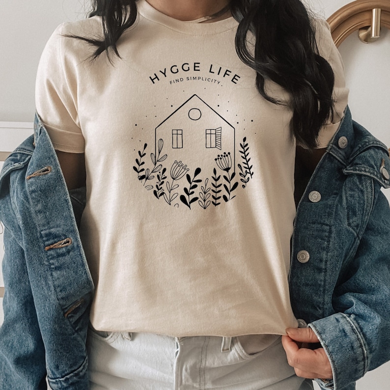 May include: A beige t-shirt featuring a black graphic design of a house with windows and a circle in the roof, surrounded by flowers and plants. The text "HYGGE LIFE FIND SIMPLICITY" is above the house.