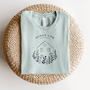 May include: A light blue t-shirt featuring a black graphic design of a house with windows and a door surrounded by flowers and the text "Hygge Life Find Simplicity".
