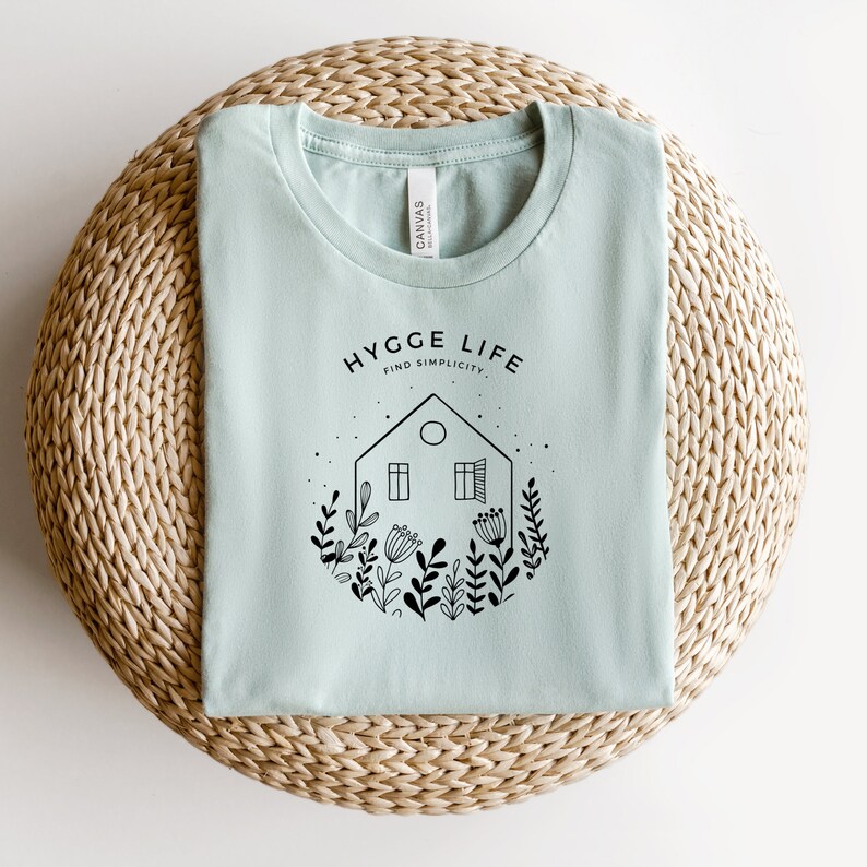 May include: A light blue t-shirt featuring a black graphic design of a house with windows and a door surrounded by flowers and the text "Hygge Life Find Simplicity".