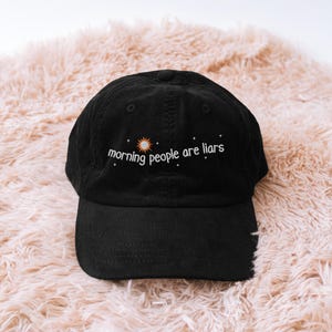 May include: Black corduroy baseball cap with the embroidered text "morning people are liars" in white, with a sun and star design. The cap has a curved brim and a fabric strap closure.