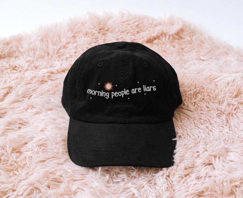 May include: Black corduroy baseball cap with the embroidered text "morning people are liars" in white, with a sun and star design. The cap has a curved brim and a fabric strap closure.