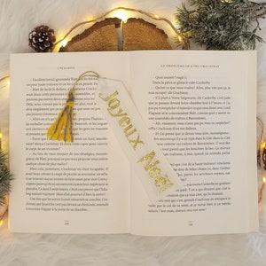 May include: A white bookmark with gold glitter lettering that says "Joyeux Noel" and a yellow tassel. The bookmark is laying open on a book with text in French.