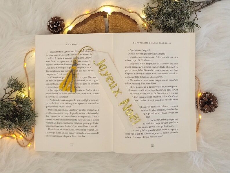 May include: A white bookmark with gold glitter lettering that says "Joyeux Noel" and a yellow tassel. The bookmark is laying open on a book with text in French.