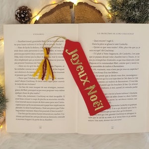 May include: A red bookmark with gold lettering that reads "Joyeux Noel" which translates to "Merry Christmas" in English. The bookmark is decorated with a gold tassel.