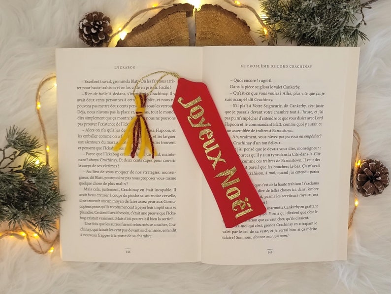 May include: A red bookmark with gold lettering that reads "Joyeux Noel" which translates to "Merry Christmas" in English. The bookmark is decorated with a gold tassel.