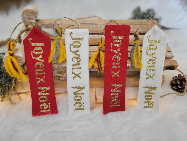 May include: Four fabric Christmas ornaments with gold glitter lettering that reads "Joyeux Noel". Two ornaments are red and two are white. Each ornament has a gold and gray tassel.