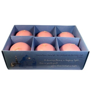 May include: A blue box containing six pink, round candles, each with a wick. The box has a decorative illustration and text that reads "A dancing flame, a leaping light..." The candles are for decoration.