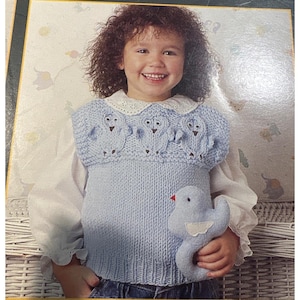 Puede incluir: Un chaleco de punto sin mangas de color azul claro con un patrón texturizado de tres pequeños pájaros en el pecho, usado sobre una camisa blanca de manga larga con cuello de encaje. Se sostiene un juguete de peluche de pájaro azul claro a juego.