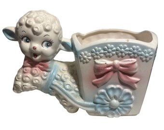 May include: A vintage ceramic planter in the shape of a lamb pulling a cart. The lamb is white with blue accents and a pink bow tie. The cart is light blue and white with pink and blue floral details.