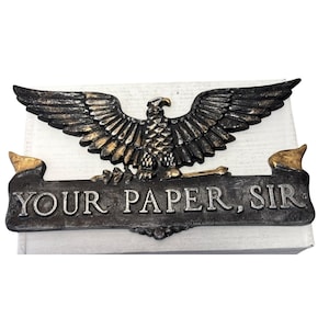 May include: A decorative metal sign featuring a detailed eagle with outstretched wings, in shades of black and gold. Below the eagle is a banner with the text "YOUR PAPER, SIR" in capital letters. The sign is mounted on a white surface.
