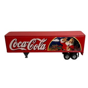 Puede incluir: Un remolque de juguete rojo de Coca-Cola con el logotipo de Coca-Cola en blanco. El remolque presenta una ilustración de Papá Noel sosteniendo una botella de Coca-Cola. El juguete tiene ruedas negras y un enganche.