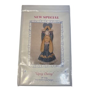 May include: A sewing pattern for an "Upsy Daisy" door knob or peg hanger. The pattern features a doll with a bunny head, wearing a blue dress. The text "SEW SPECIAL" is at the top. The hanger is 29 inches long.